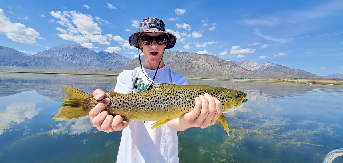 A smiling fly fisherman holding a rainbow trout on a lake in a boat.