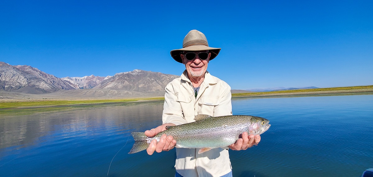 A smiling fly fisherman holding a rainbow trout on a lake in a boat.