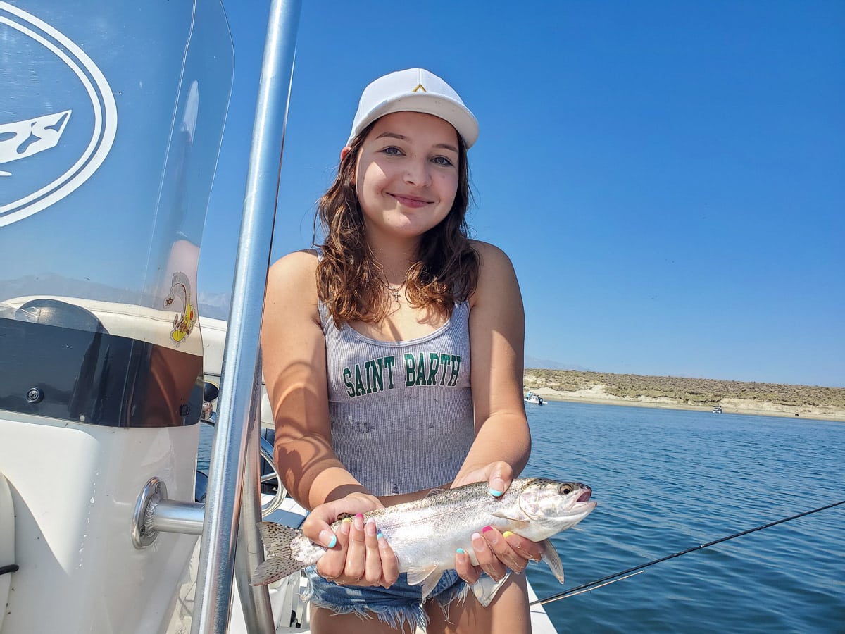 A smiling young female fly fisherman holding a rainbow trout on a lake in a boat.