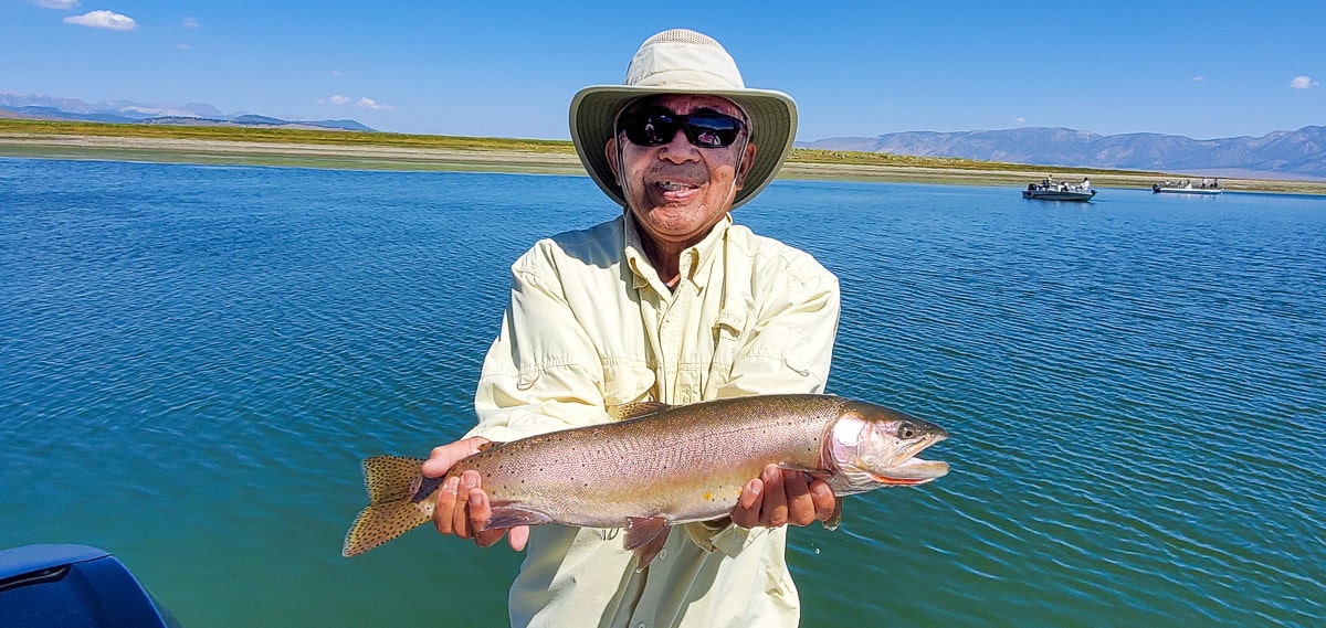 A smiling fly fisherman holding a cutthroat trout on a lake in a boat.