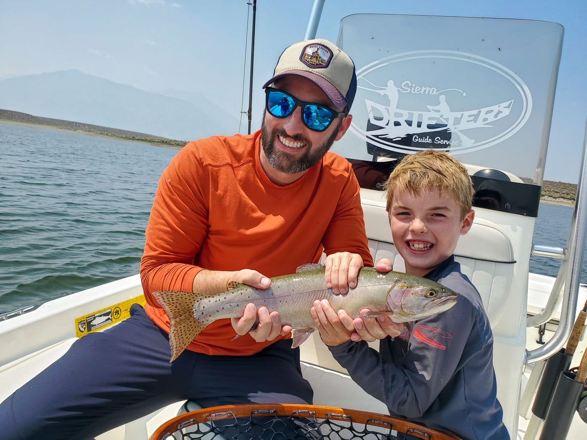 A smiling fly fisherman and a young boy holding a cutthroat trout on a lake in a boat.