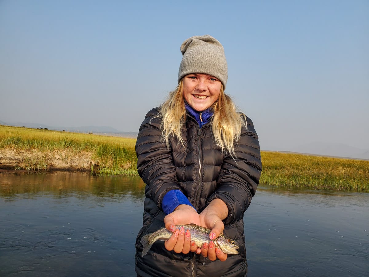 A young girl holding a rainbow trout while standing in a river with tall green grass on the banks.