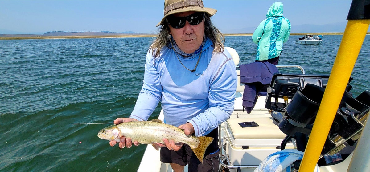 A smiling fly fisherman holding a cutthroat trout on a lake in a boat.