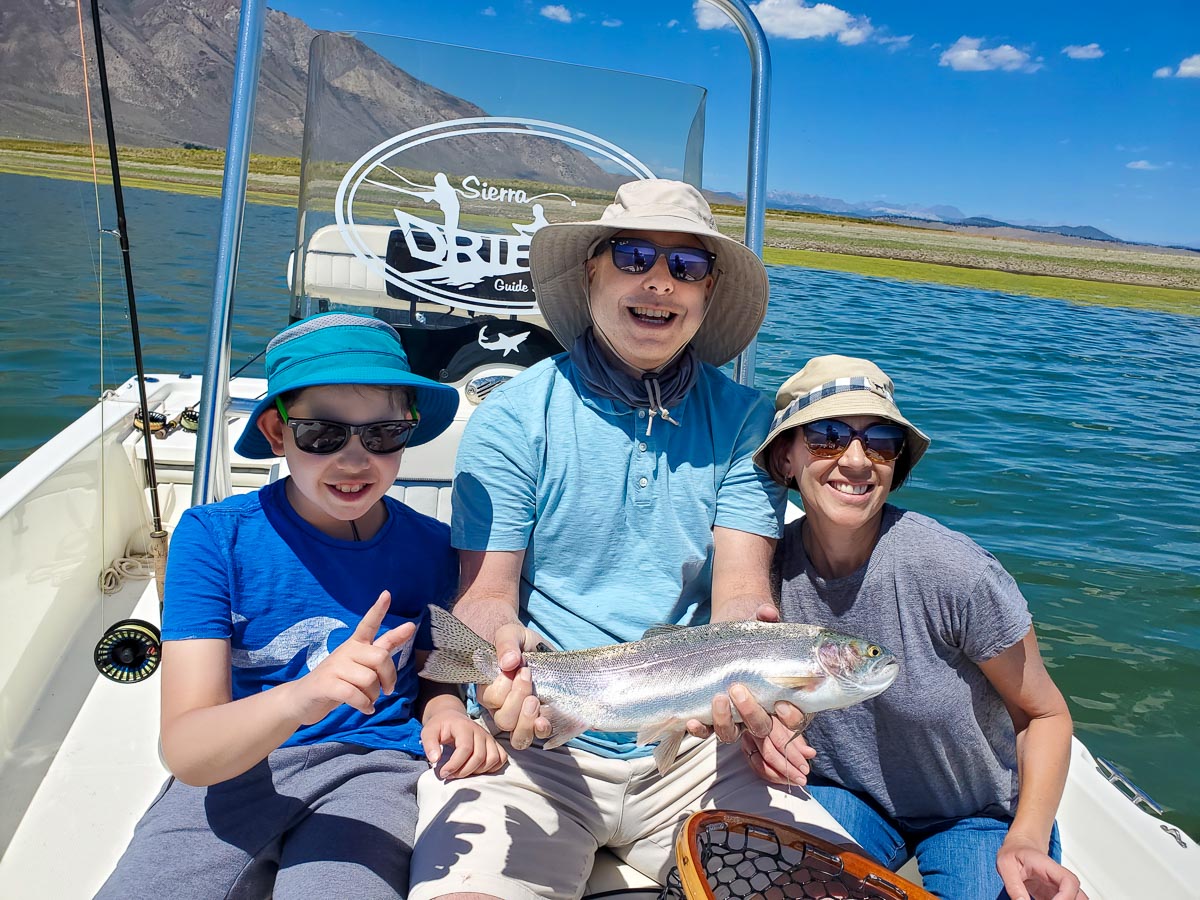 A family (man, woman, and young boy) of anglers holding a rainbow trout in a boat on a lake.