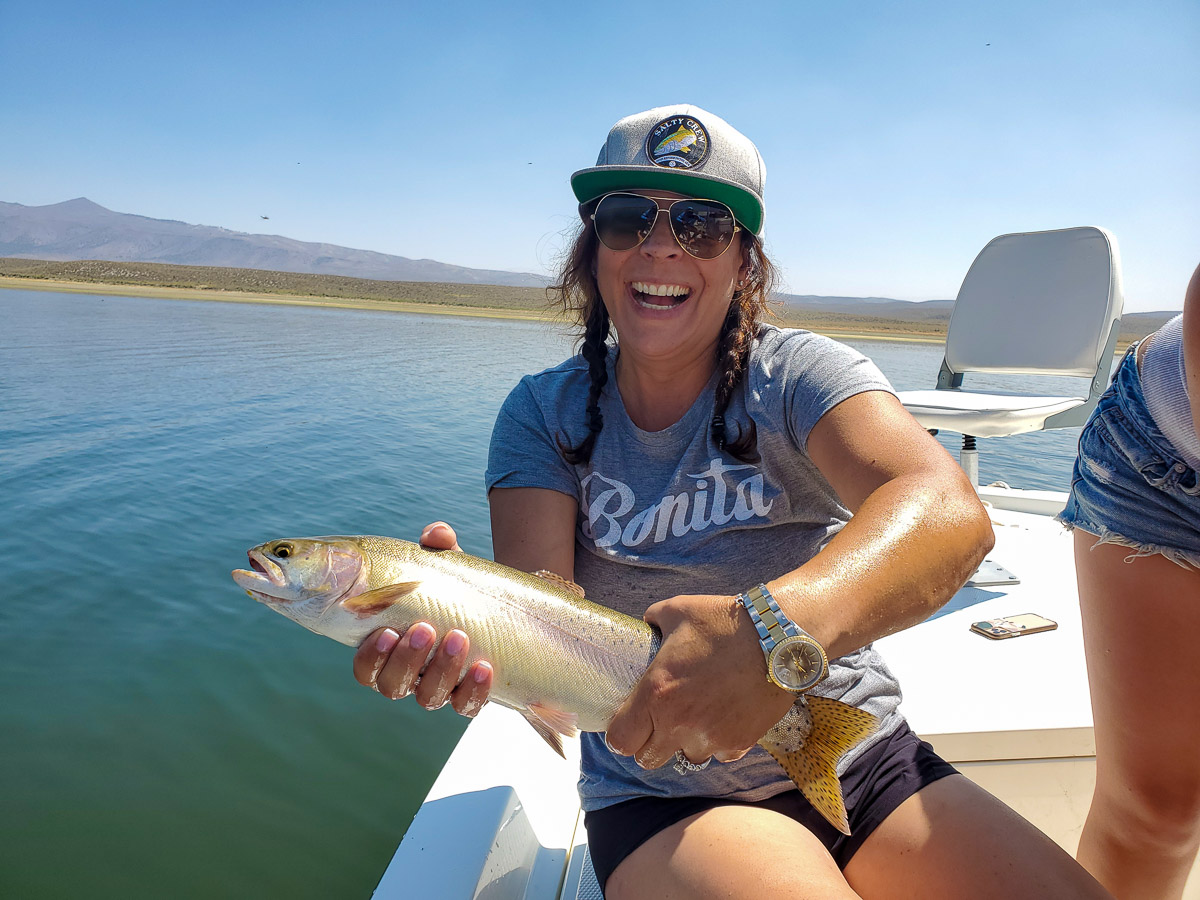 A female fly angler holding a large cutthroat trout in a boat on a glassy surfaced lake.