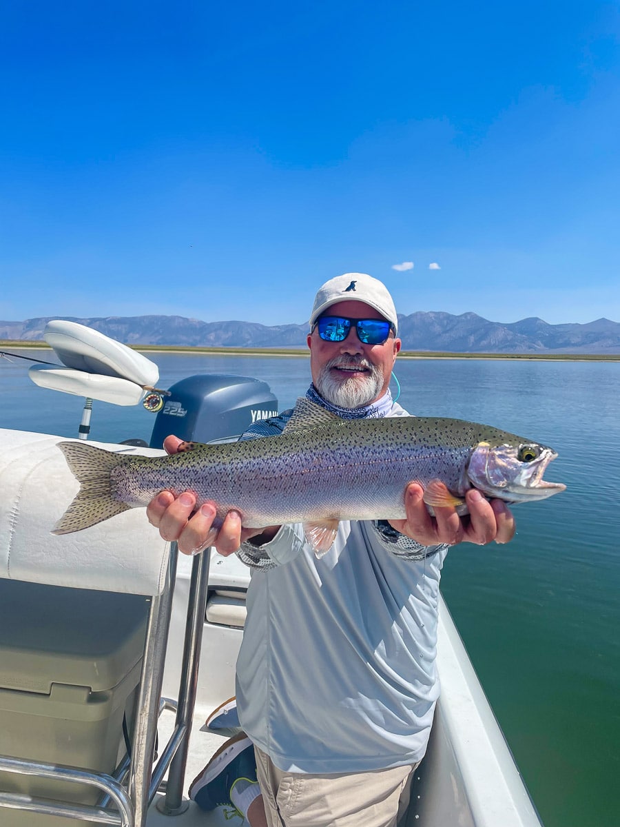 A smiling fly fisherman holding a rainbow trout on a lake in a boat.