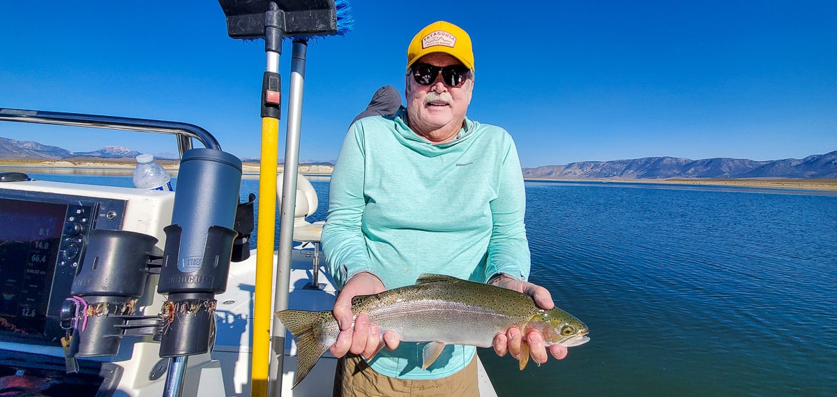 A smiling fly fisherman holding a rainbow trout on a lake in a boat.