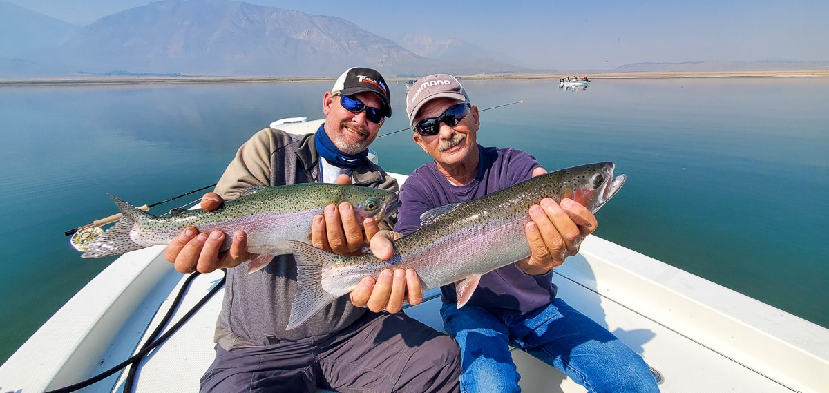 A pair of smiling fly fisherman holding a pair of rainbow trout on a lake in a boat.