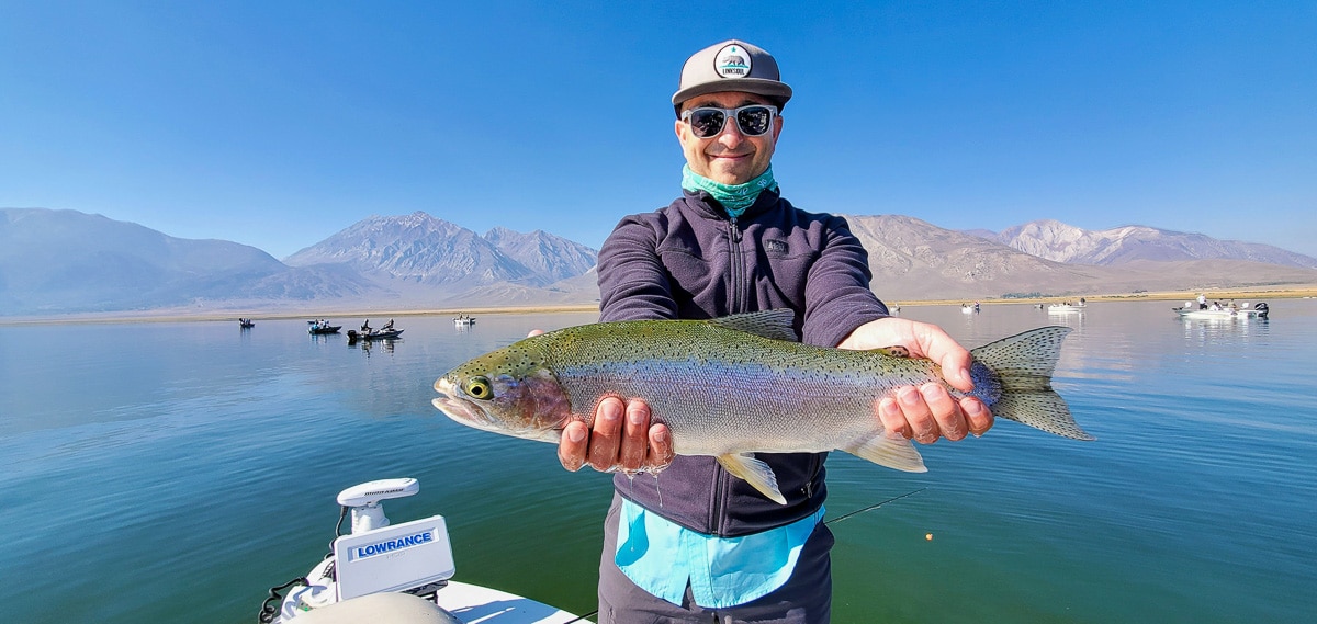 A smiling fly fisherman holding a rainbow trout on a lake in a boat.