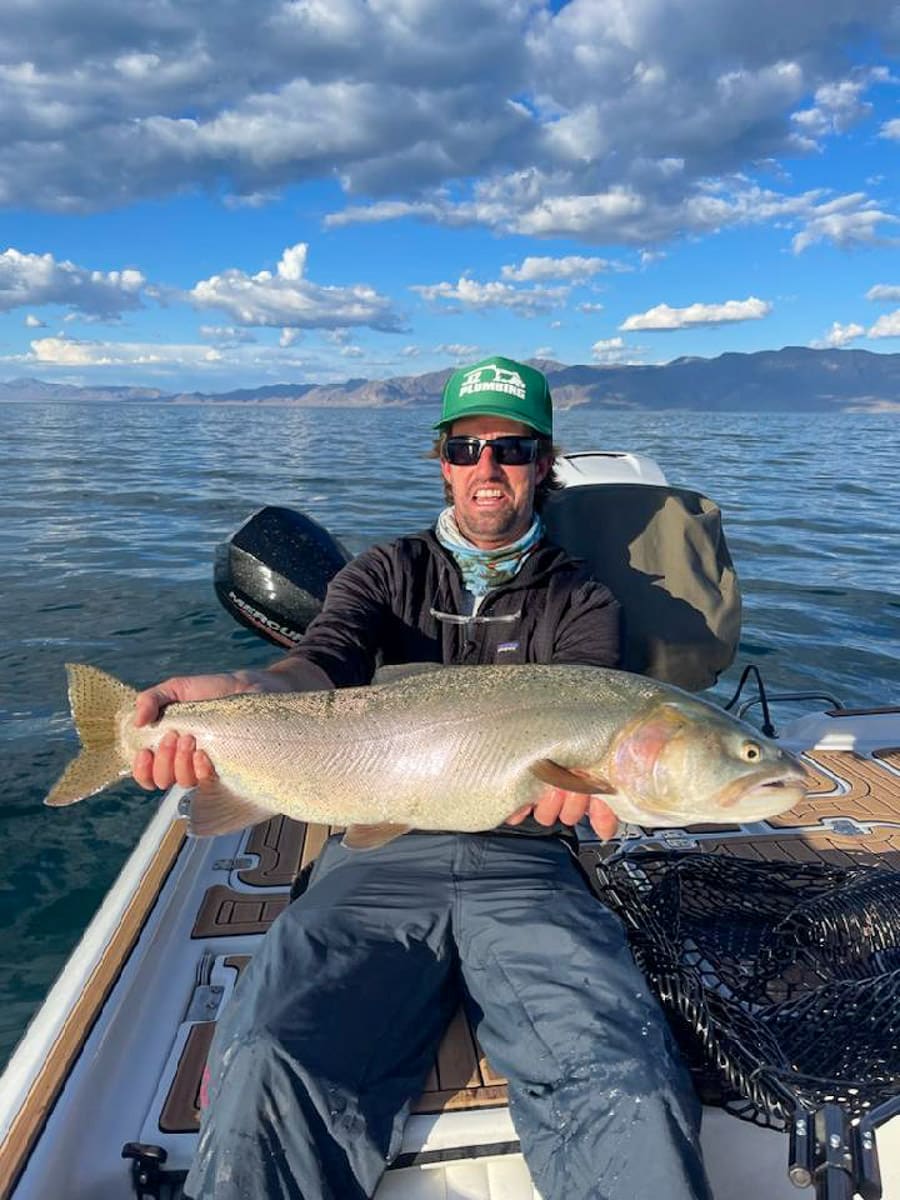 A fly fisherman on a lake in a boat holding a behemoth cutthroat trout.