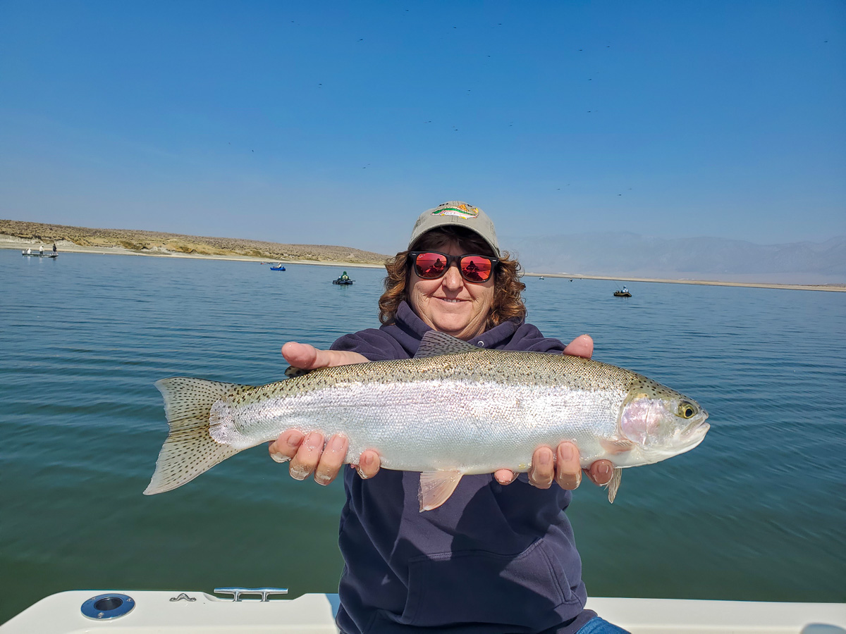 A smiling fly fisherwomman holding a rainbow trout on a lake in a boat.