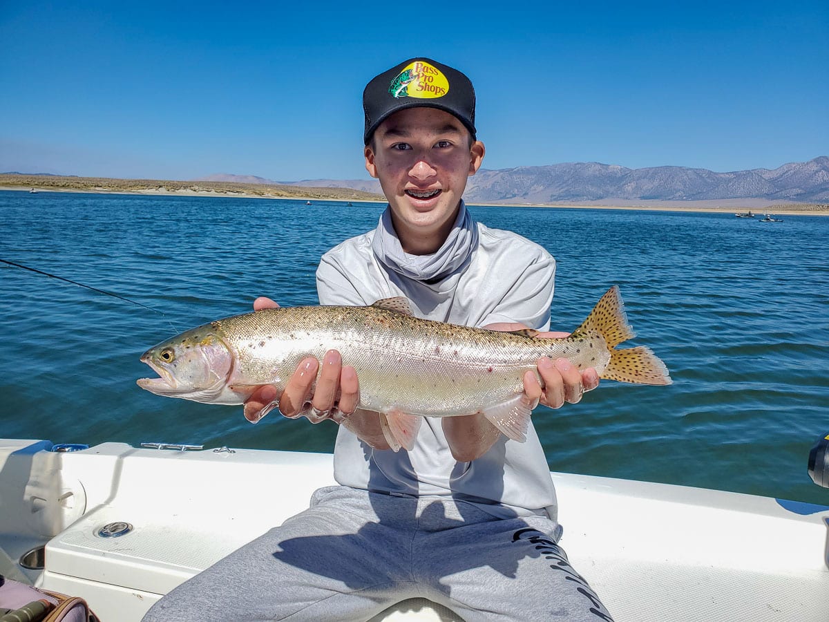 A smiling fly fisherman holding a rainbow trout on a lake in a boat.