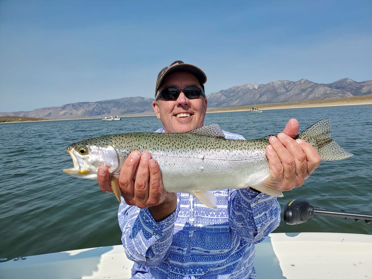 A smiling fly fisherman holding a rainbow trout on a lake in a boat.