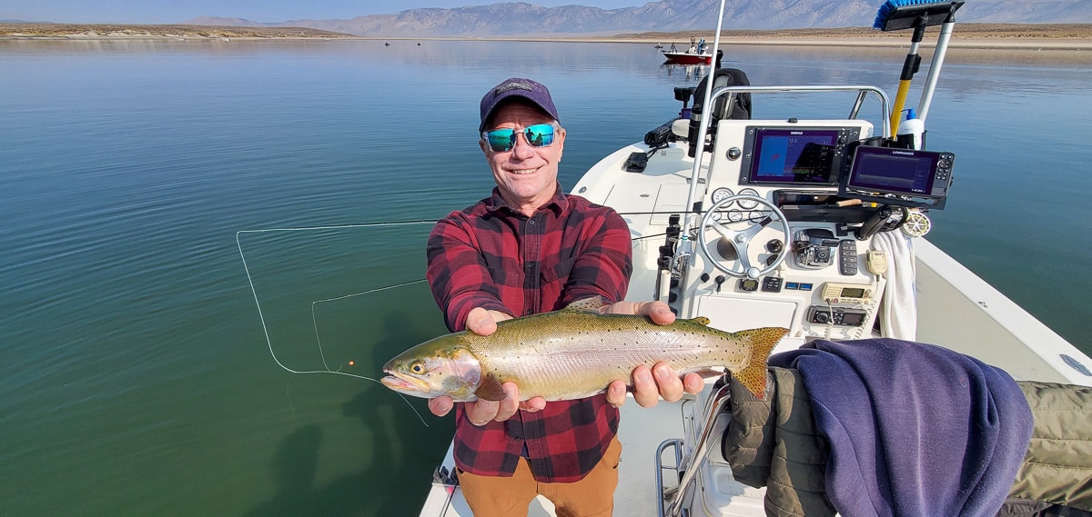 A smiling fly fisherman holding a cutthroat trout on a lake in a boat.