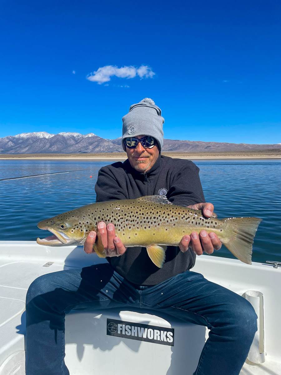 A smiling fly fisherman holding a brown trout on a lake in a boat.