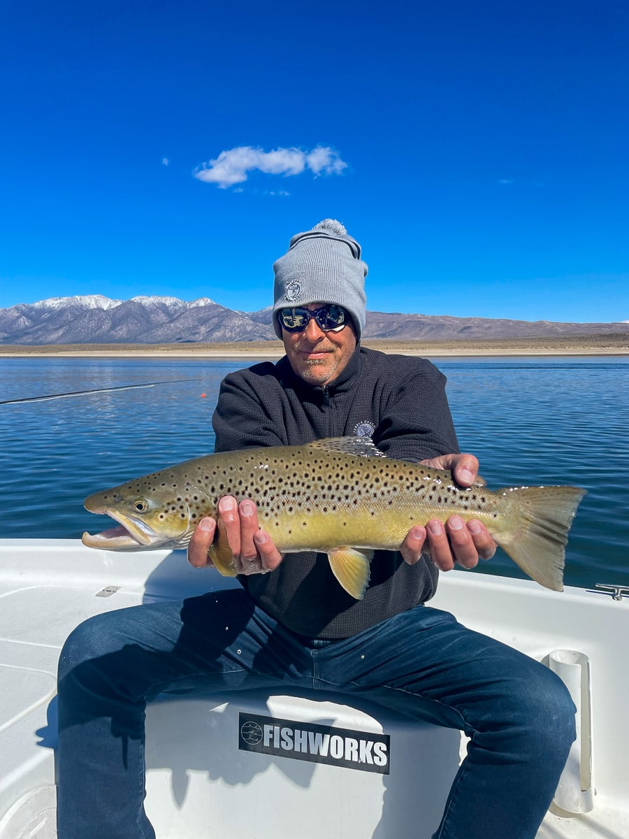 A smiling fly fisherman holding a brown trout on a lake in a boat.