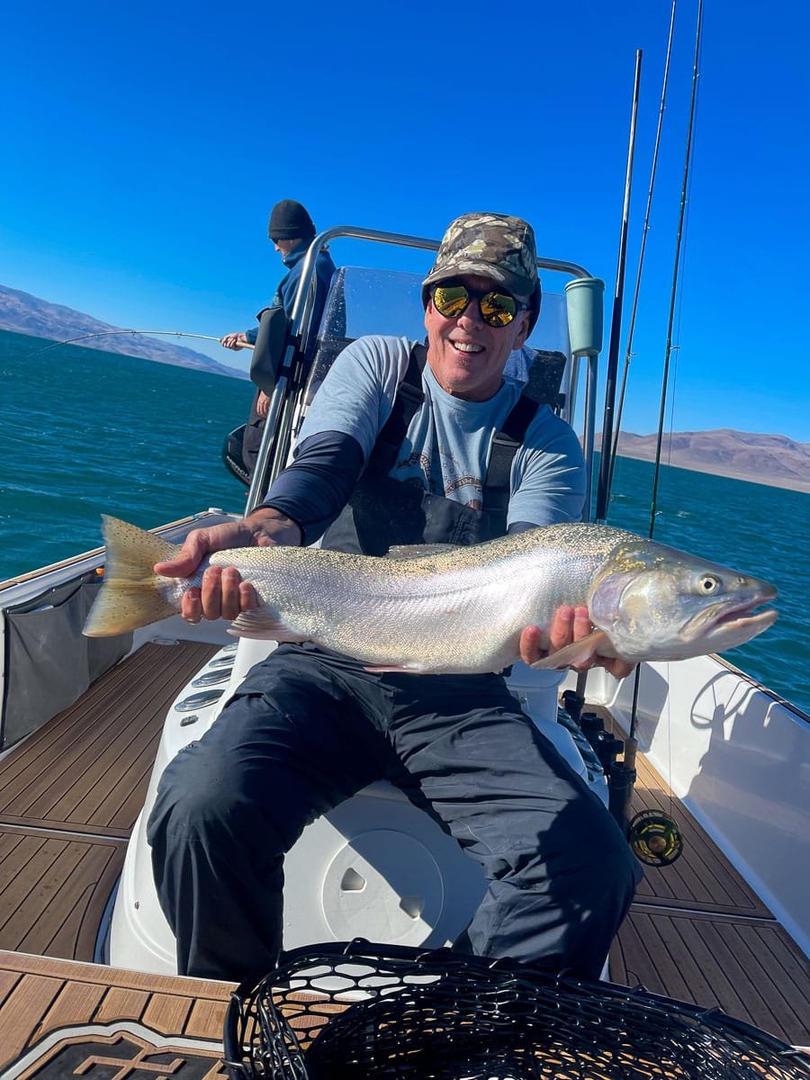 A smiling fly fisherman holding a cutthroat trout on a lake in a boat.