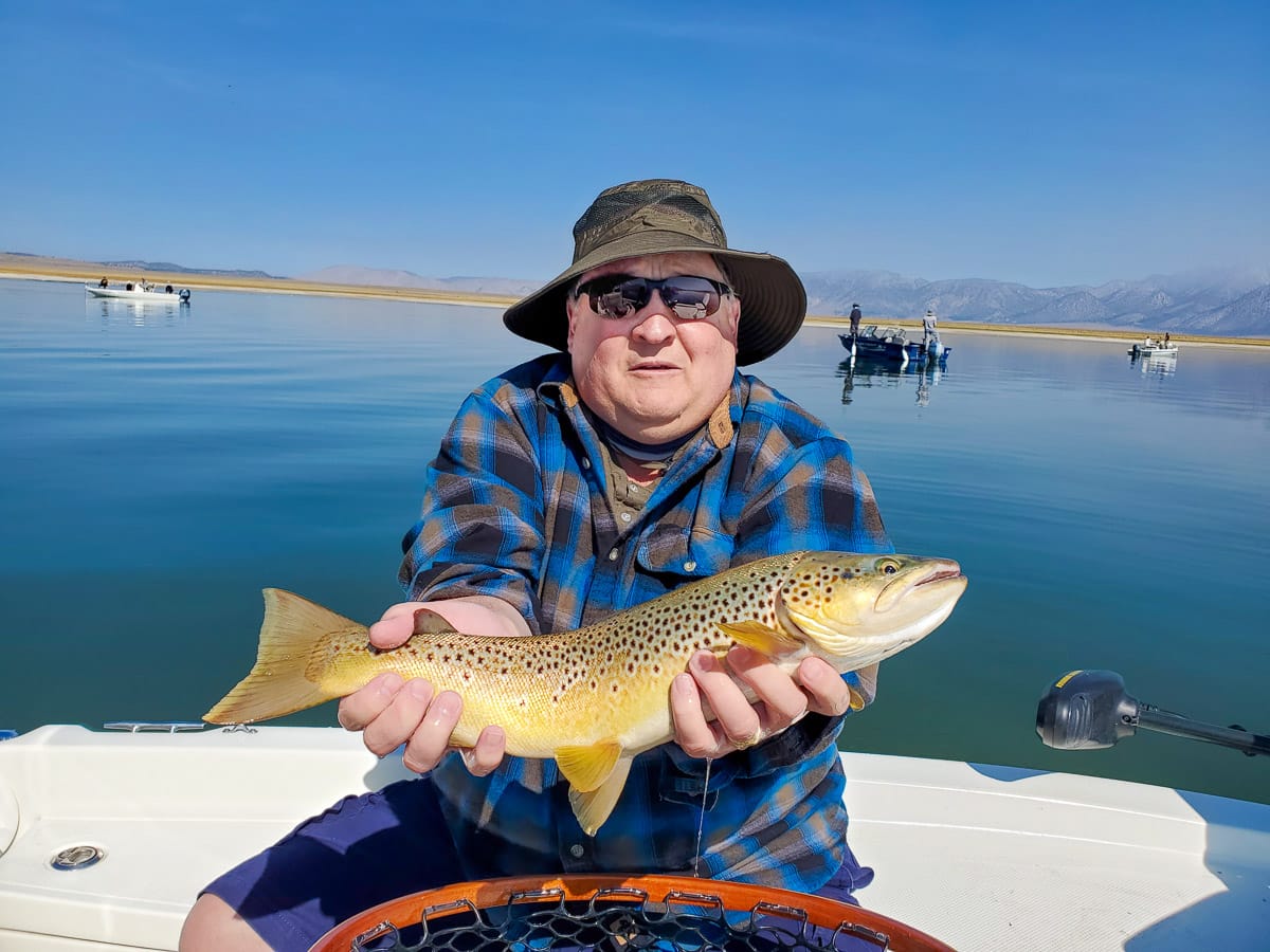 A smiling fly fisherman holding a brown trout on a lake in a boat.