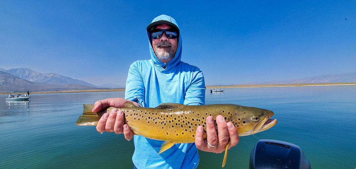 A smiling fly fisherman holding a brown trout on a lake in a boat.