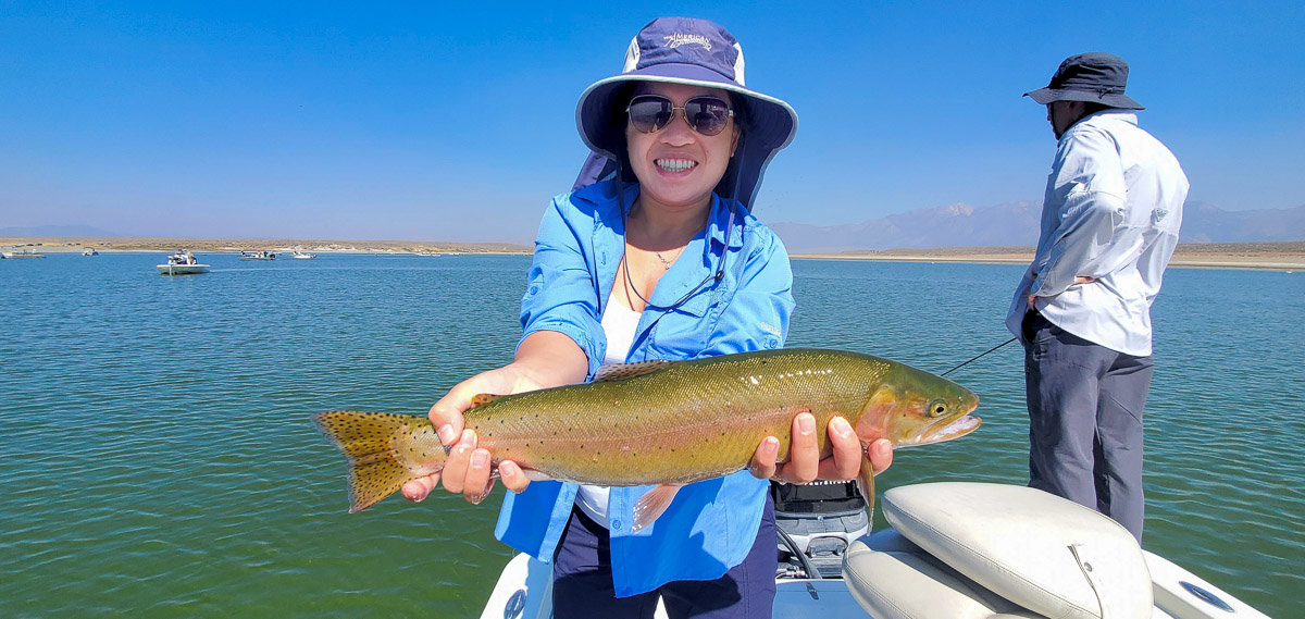 A smiling lady fly fisherman holding a cutthroat trout on a lake in a boat.