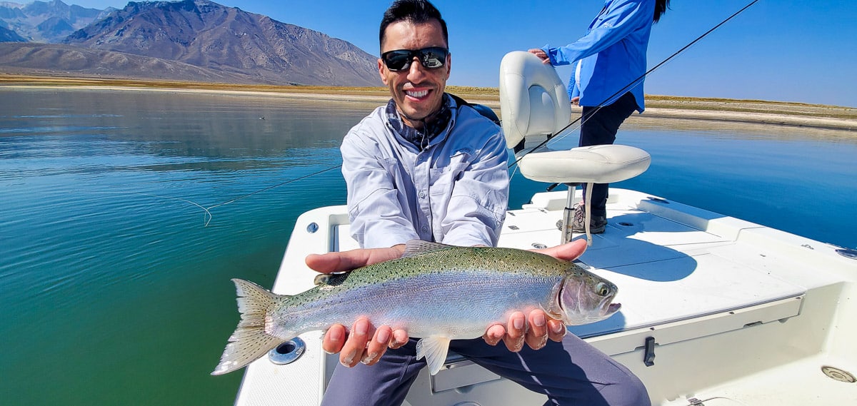 A smiling fly fisherman holding a rainbow trout on a lake in a boat.