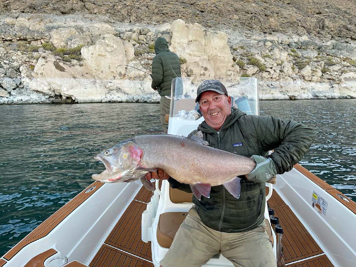 A smiling fly fisherman holding a cutthroat trout on a lake in a boat.