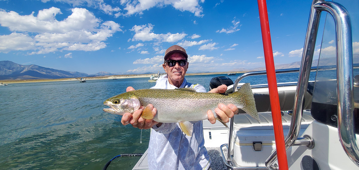 A smiling fly fisherman holding a rainbow trout on a lake in a boat.