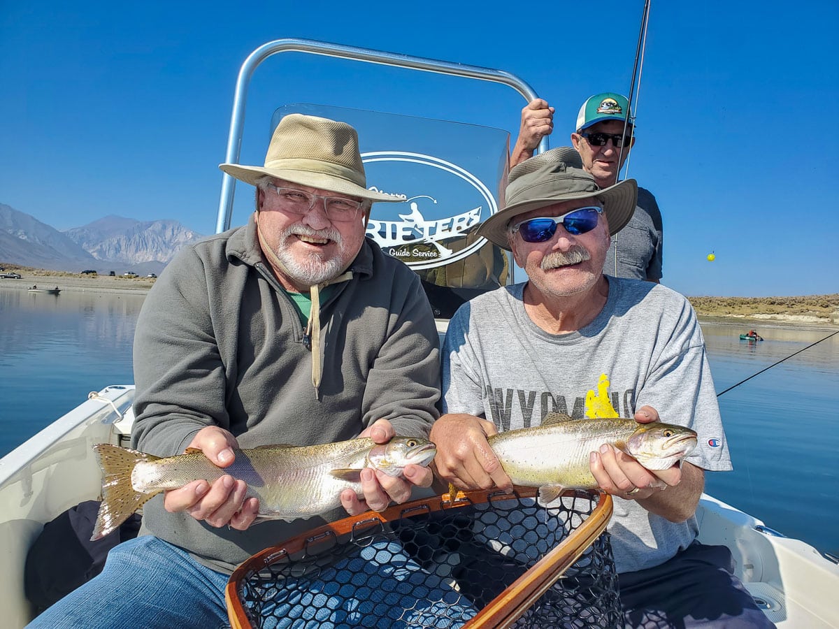 A pair of smiling fly fisherman holding 2 rainbow trout on a lake in a boat.