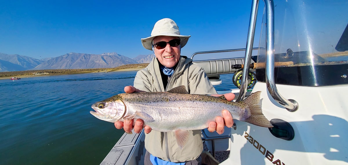 A smiling fly fisherman holding a rainbow trout on a lake in a boat.