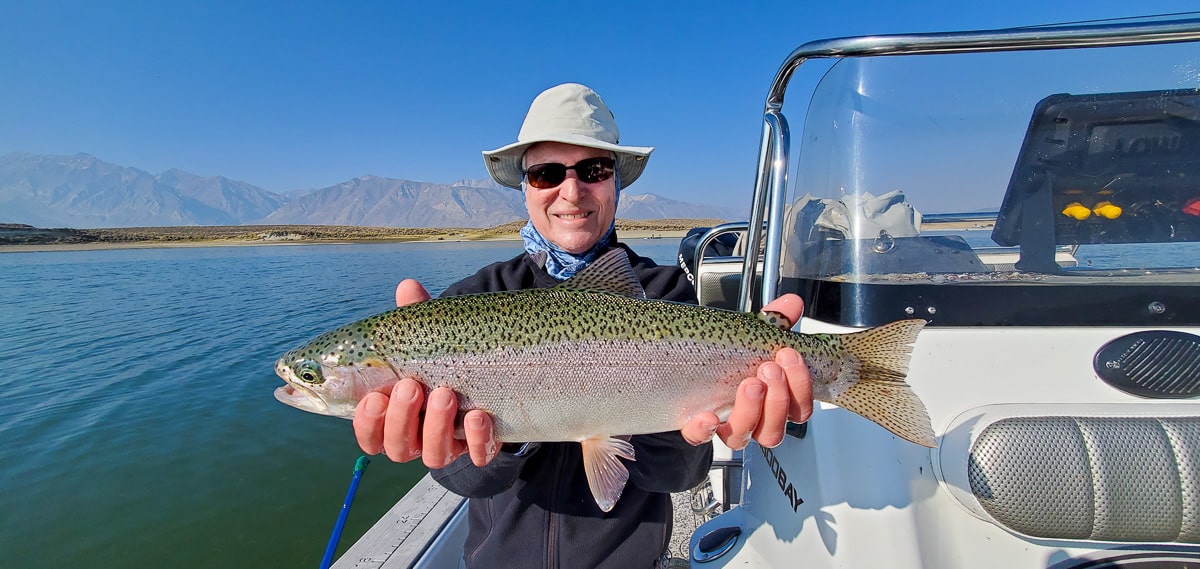 A smiling fly fisherman holding a rainbow trout on a lake in a boat.