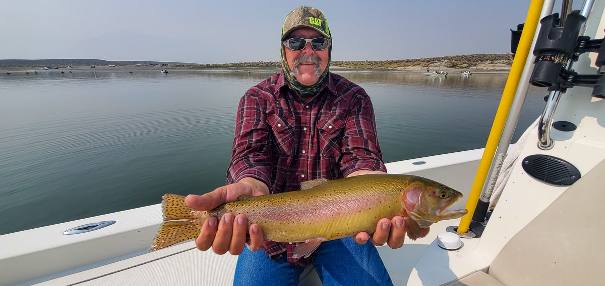 A smiling fly fisherman holding a cutthroat trout on a lake in a boat.