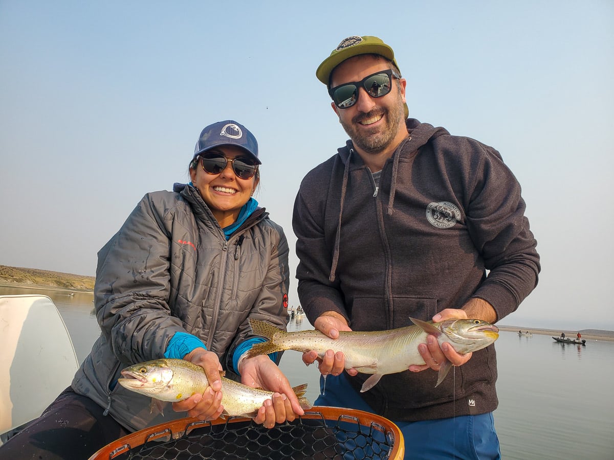 A smiling pair of fly fisherman holding a couple trout on a lake in a boat.