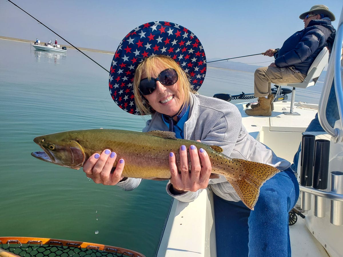 A fly fisherwoman holding a cutthroat in a boat on a lake.