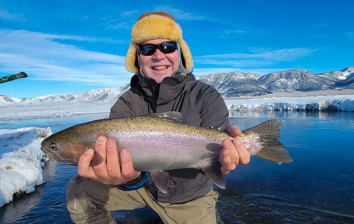 A smiling fly fisherman holding a rainbow trout on a river.