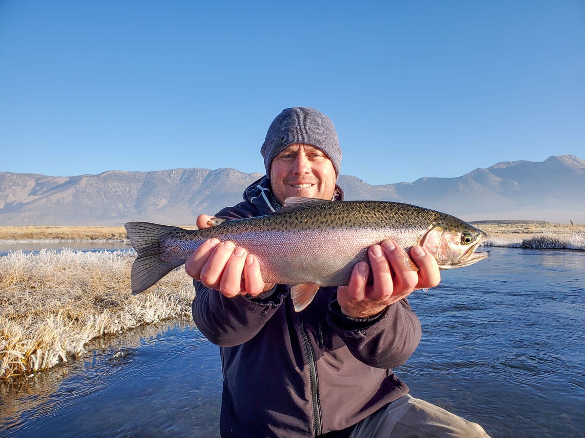 A smiling fly fisherman holding a rainbow trout on a river.