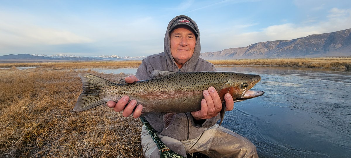 A smiling fly fisherman holding a rainbow trout on a river.