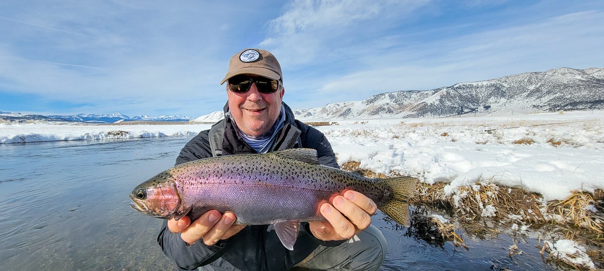 A smiling fly fisherman holding a rainbow trout on a river.