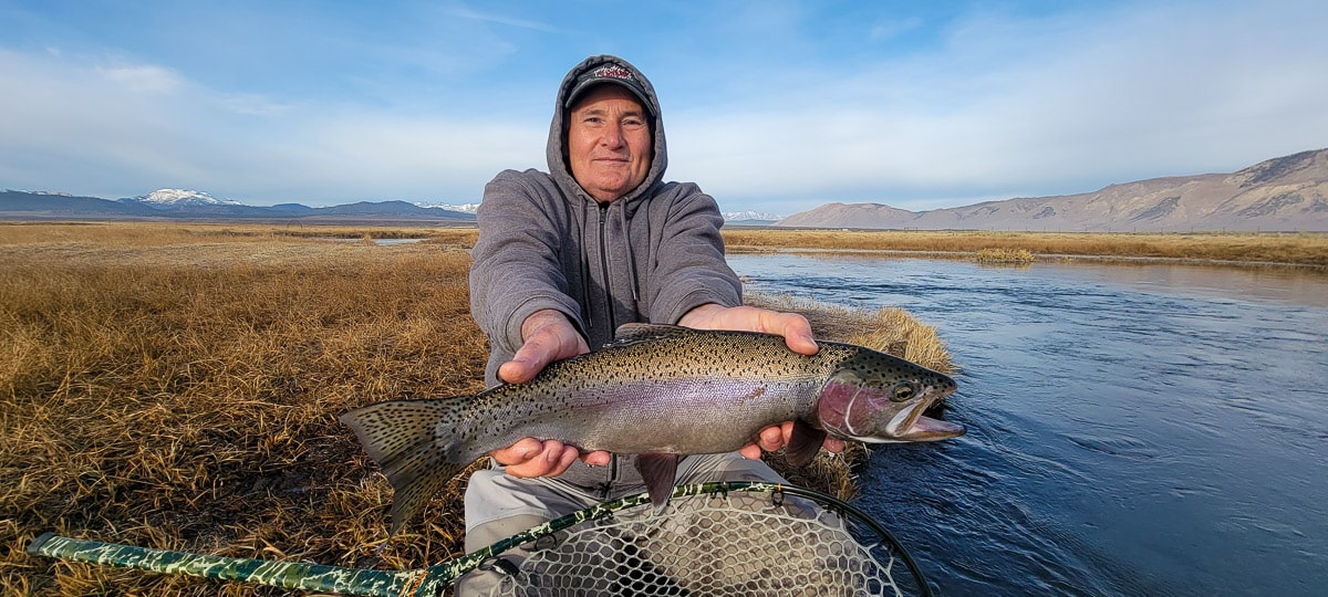 A smiling fly fisherman holding a rainbow trout on a river.