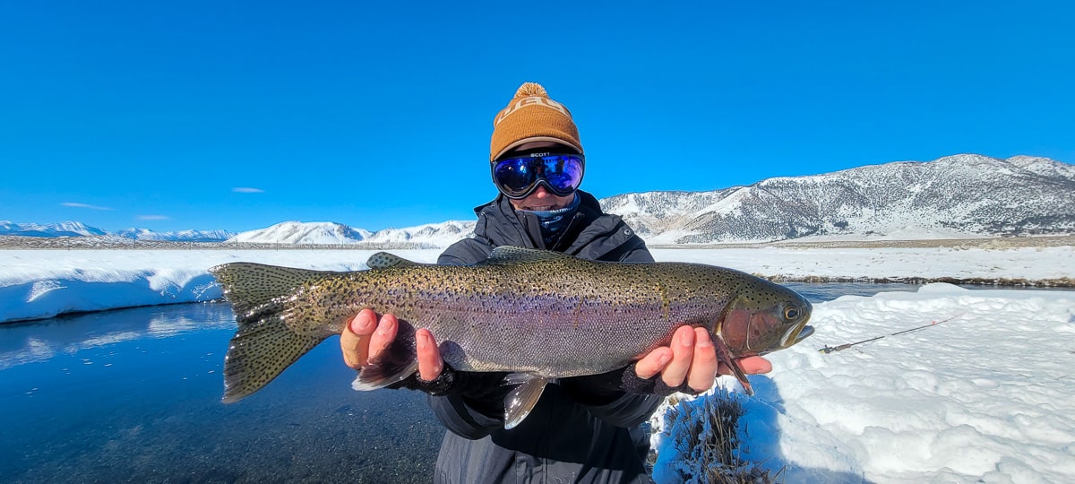 A smiling fly fisherman holding a rainbow trout on a river.