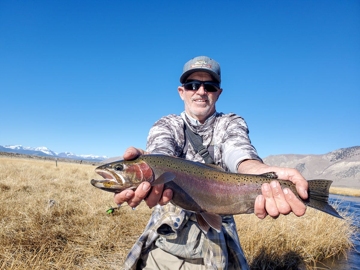 A smiling fly fisherman holding a rainbow trout on a river.