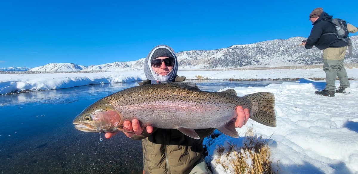 A smiling fly fisherman holding a rainbow trout on a river.