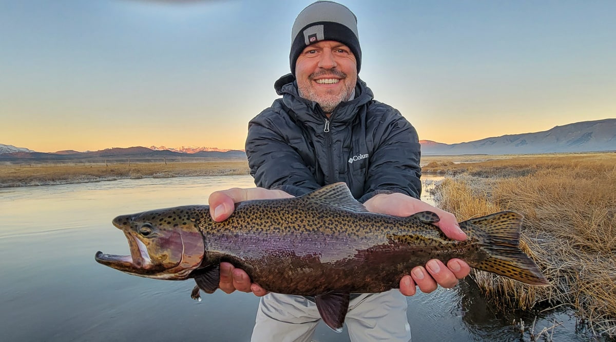 A smiling fly fisherman holding a rainbow trout on a river.