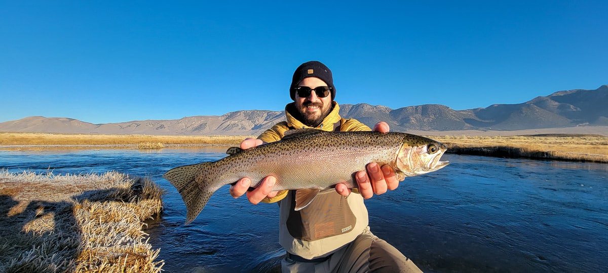 A smiling fly fisherman holding a rainbow trout on a river.