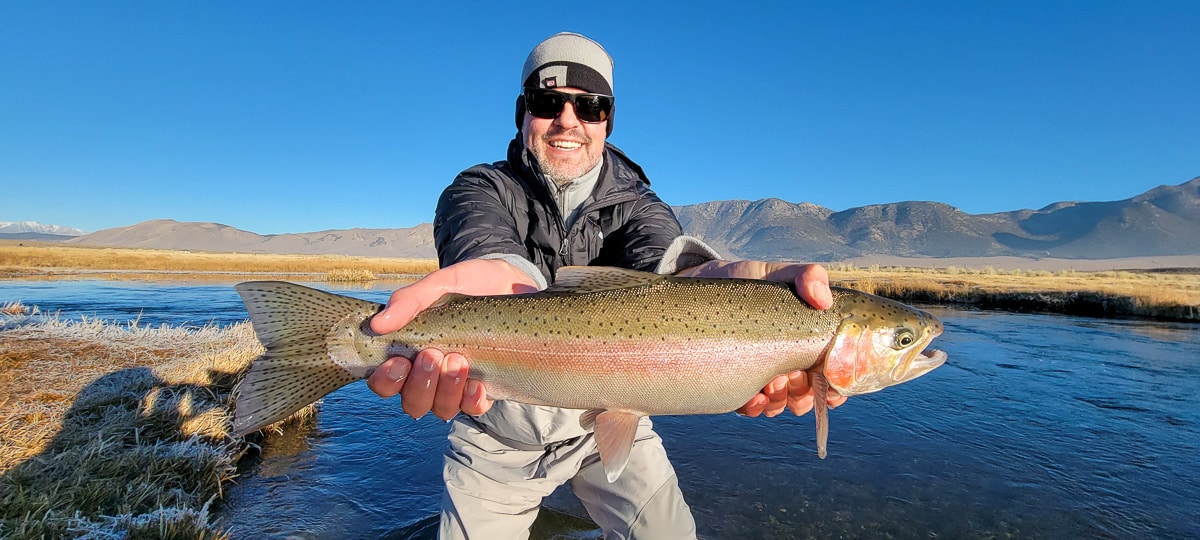 A smiling angler holding a giant rainbow trout on a river.