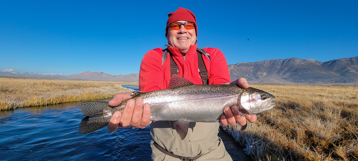 A smiling fly fisherman holding a rainbow trout on a river.