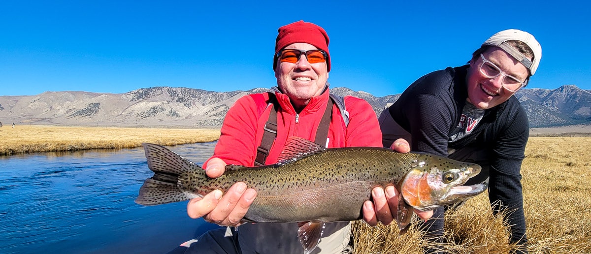 A smiling fly fisherman holding a rainbow trout on a river.