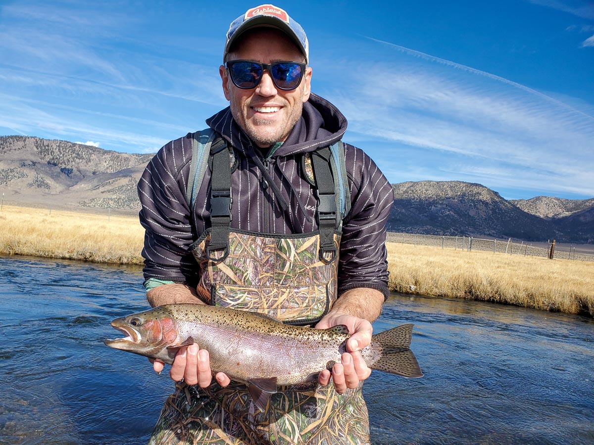 A smiling fly fisherman holding a rainbow trout on a river.