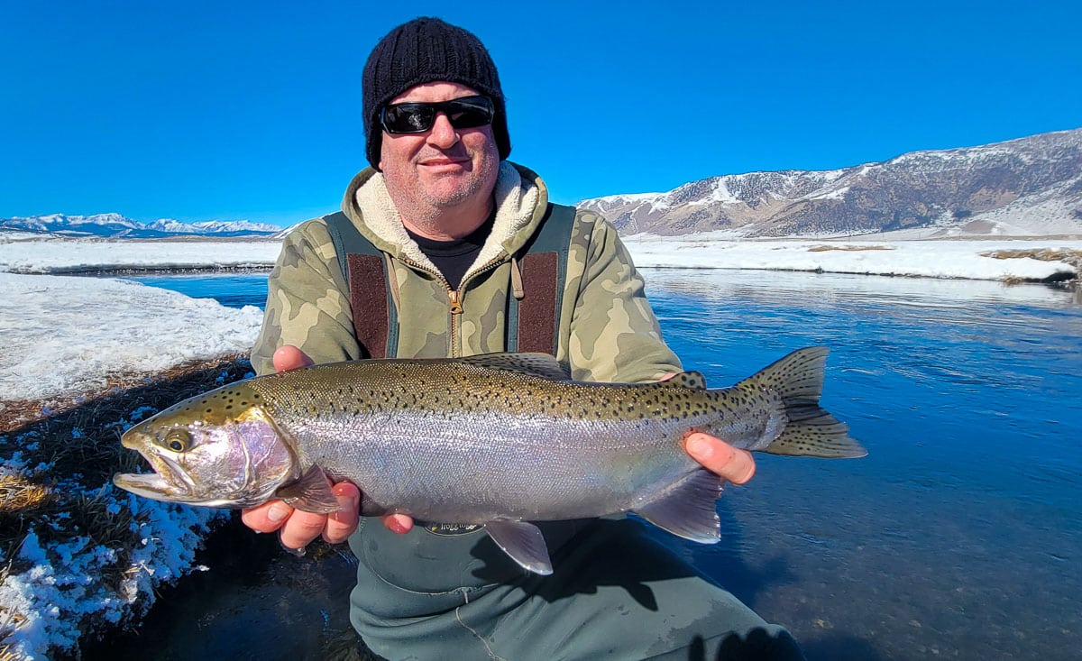 A smiling fly fisherman holding a rainbow trout on a river.