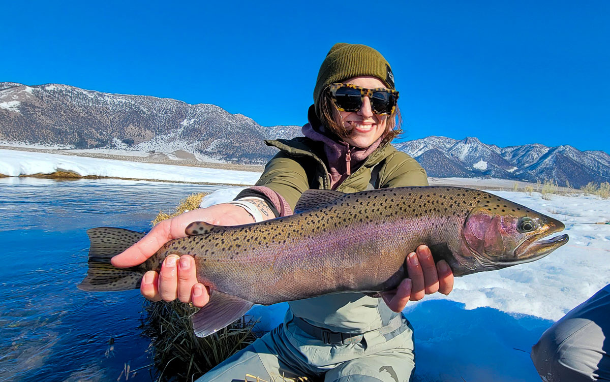 A smiling female angler holding a giant rainbow trout on a river.