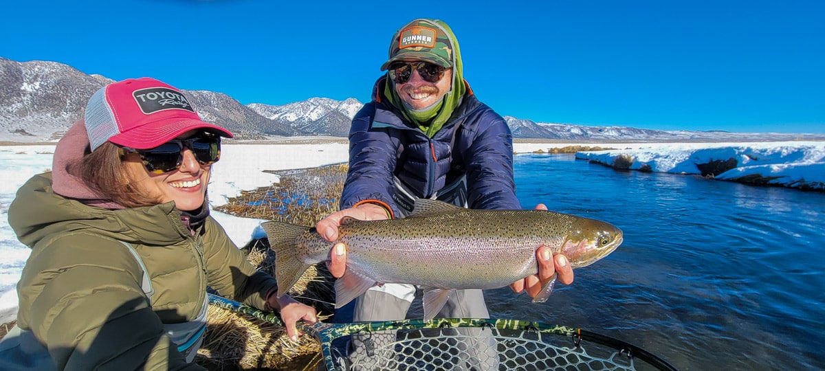 A smiling angler holding a giant rainbow trout on a river with a young lady in a red hat holding a net under the fish.